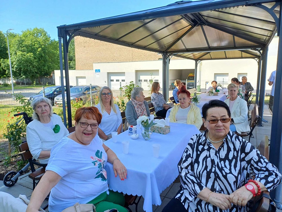 Seniorki z DDS i goście siedzą przy stole, przykryty białym obrusem, w tle zieleń 