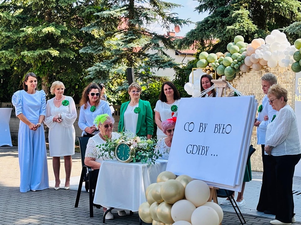Kadra zarządzająca DDS stoi, przed nimi przy stoliku siedzą dwie seniorki, po prawej duża biała kartka z napisem Co by było gdyby... w tle zieolne drzewa