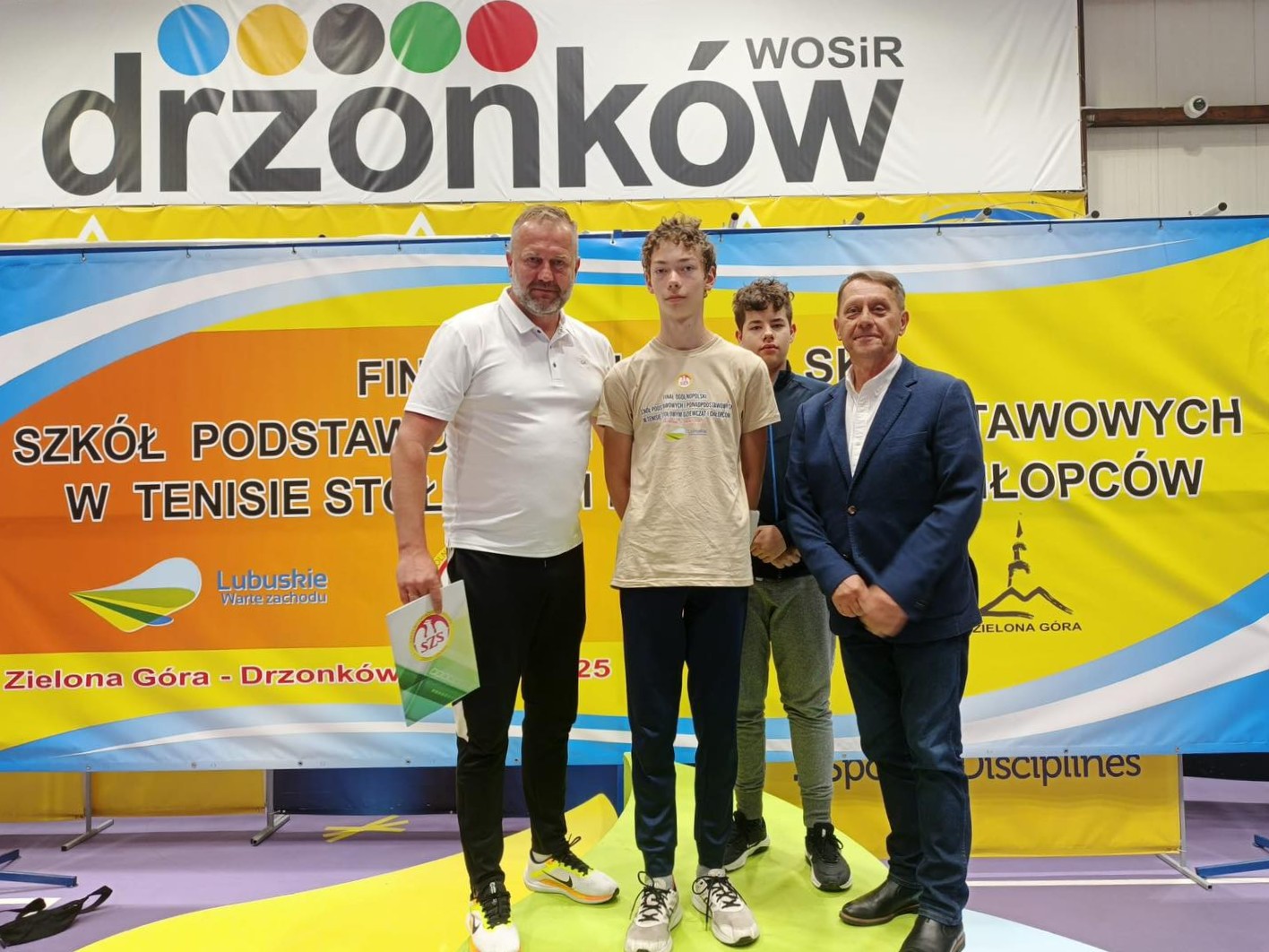 Na podeście stoją nauczyciel (od lewej) i dwóch uczniów i mężczyzna. W tle baner z napisem: Dżonków WOSiR Finał Ogólnopolski Szkół podstawowych i ponadpodstawowcych w tenisie 