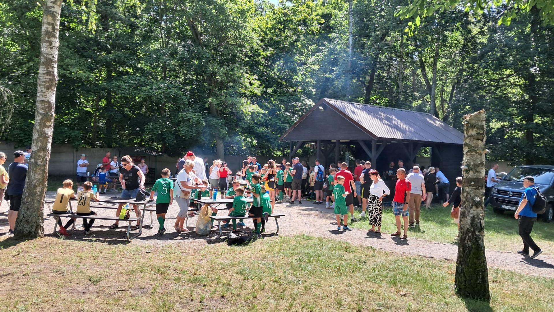 Grupa dzieci siedzi na ławkach lub stoi; w oddali wiata nad którą unosi się dym. w tle zielone drzewa