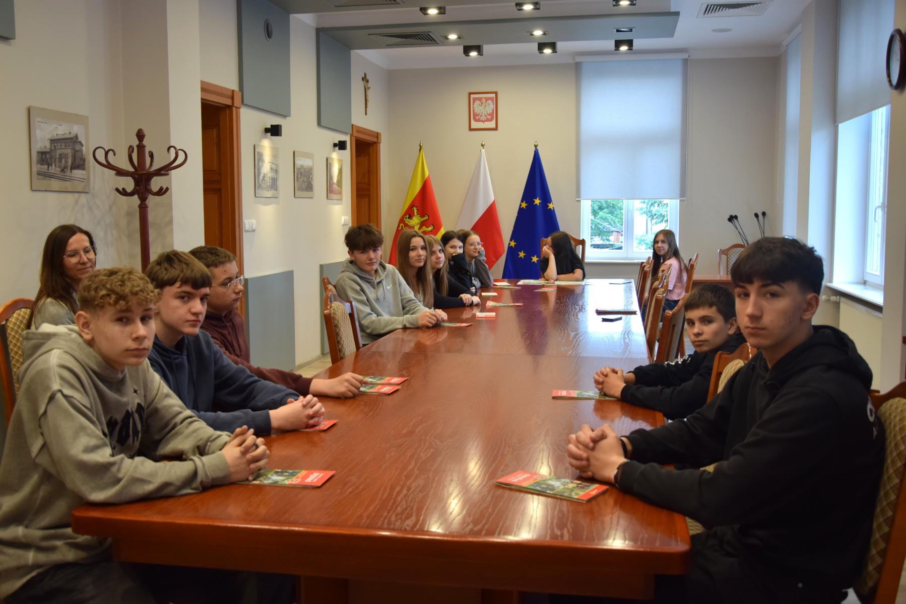Uczniwoei siedzą przy dugim stole w sali posiedzeń. w tle flagi gminna, polski i UE