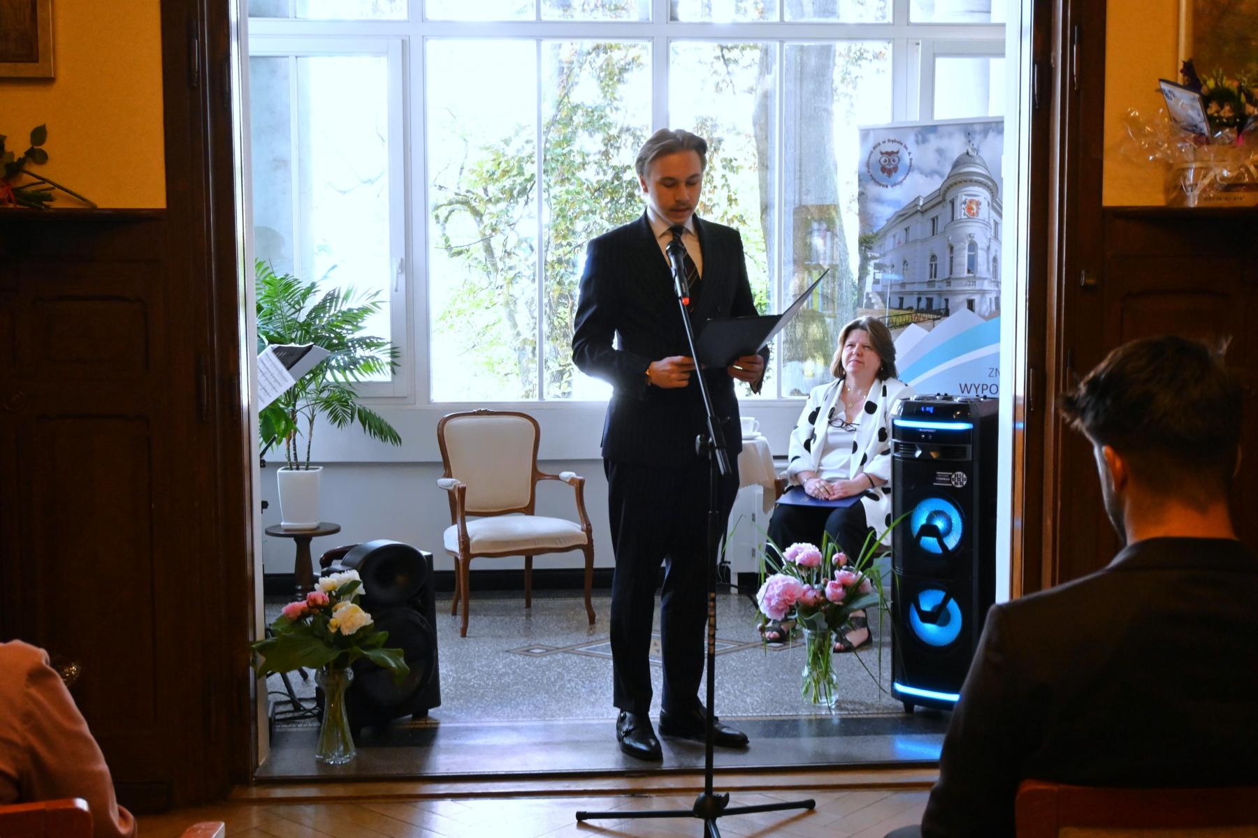 Młody mężczyzna stoi przy mikrofonie, w rękach trzyma rozłożoną teczkę, w tle po prawej stronie siedzi kobieta.