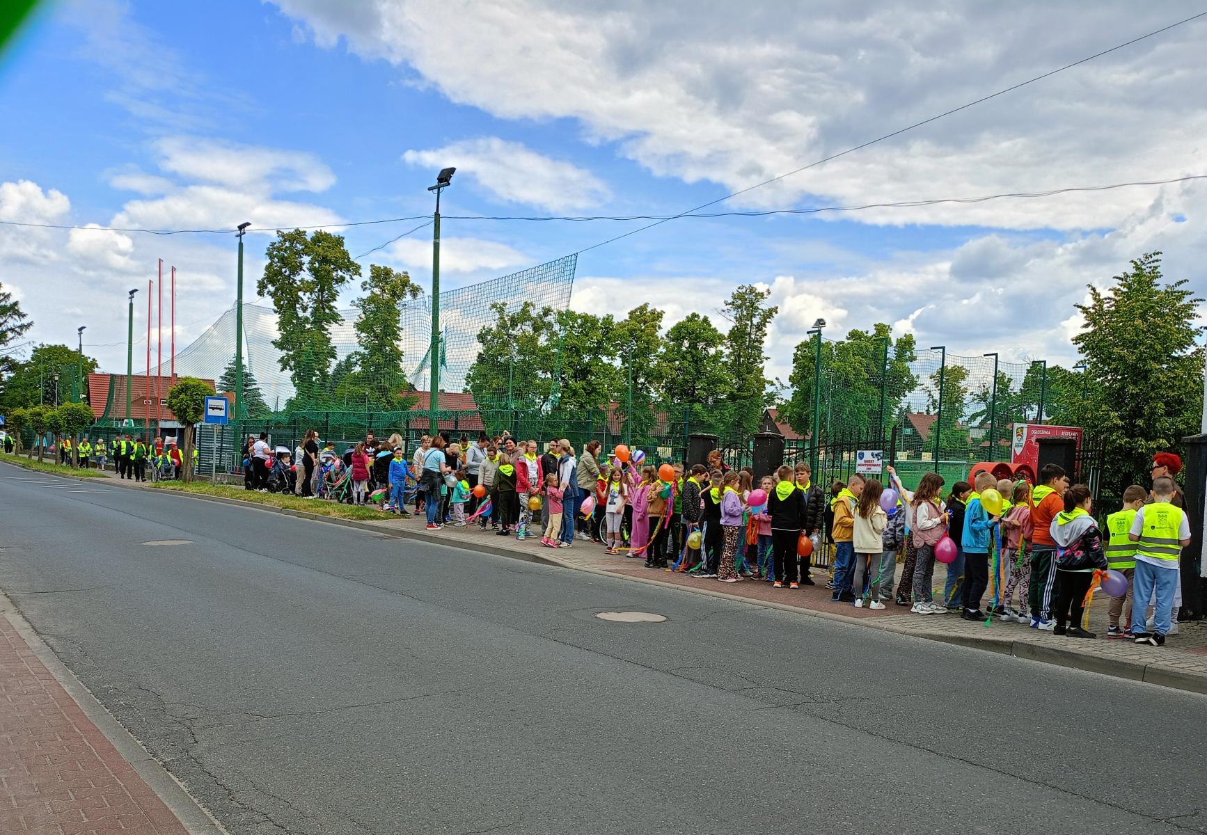 Długa kolejka dzieci i osób niepełnopsrawnych na chodniku. z przodu jezdnia, w tle dach domu, zielone drzewa, niebo z chmurami, lampy