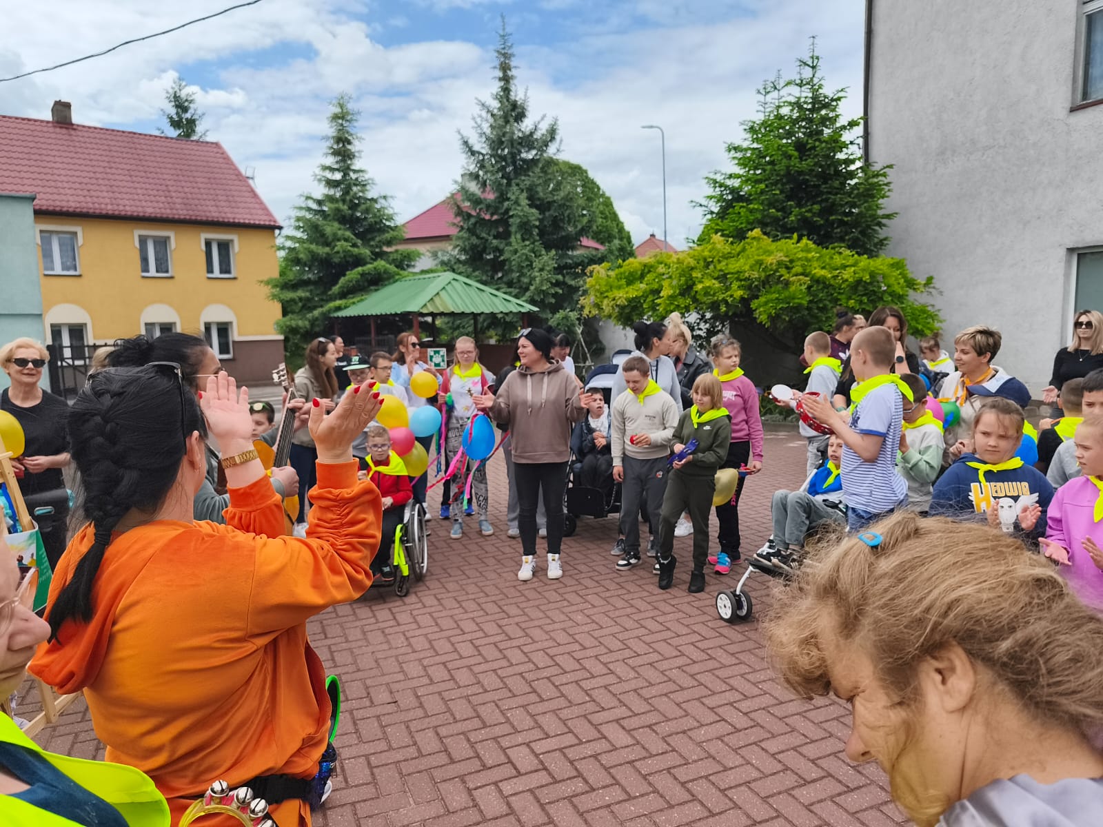 grupa ludzi stoi w okegu na placu, klaszczą, w tle budynke, zielone drzewa, niebieskie niebo 
