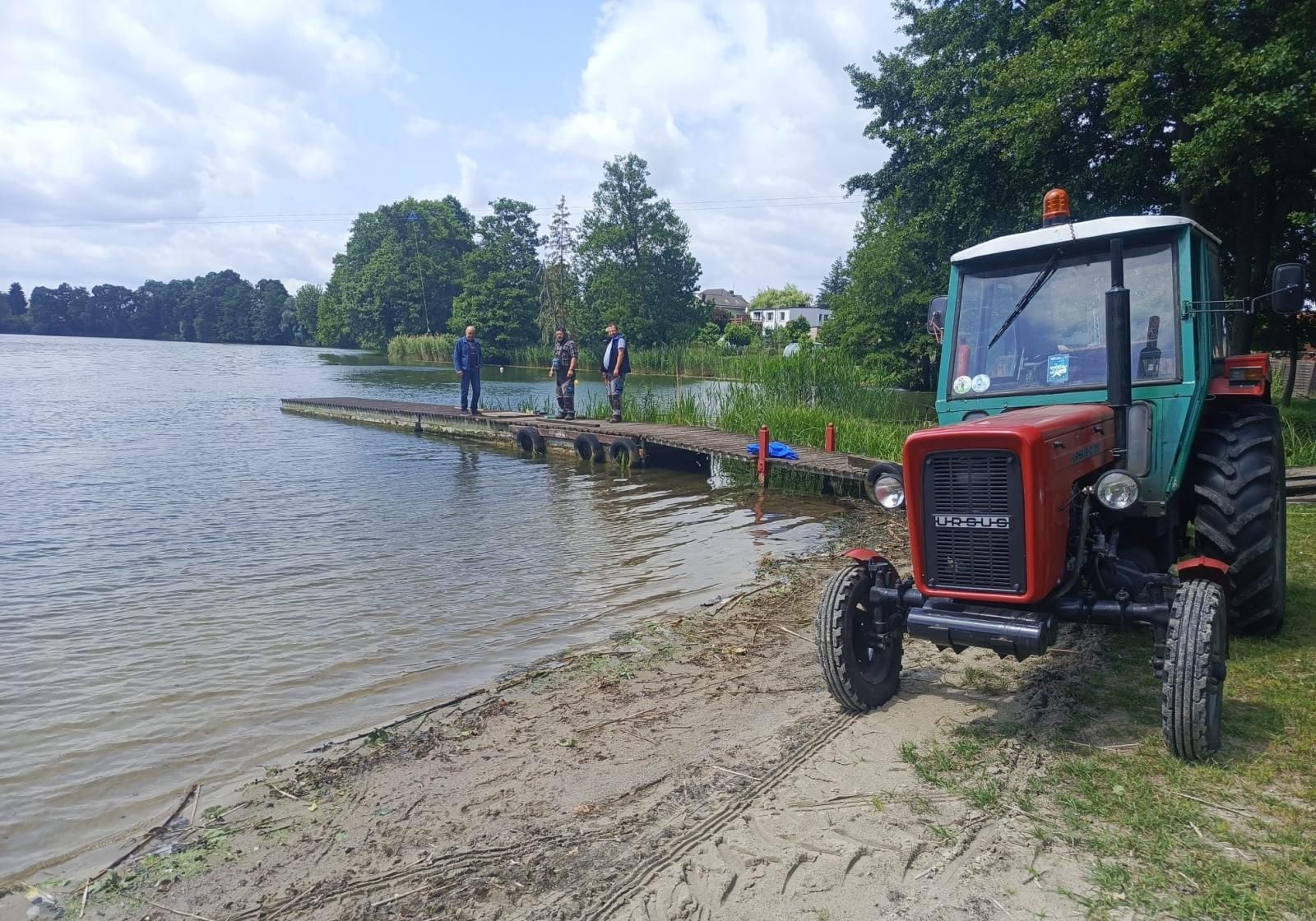 pomost, na pomoście trzy oosby stoją, traktor po prawej stronie stoi przy brzegu jeziora