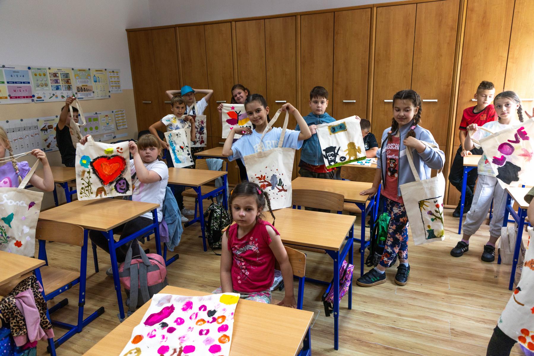 Dzieci w klasie stoją lub siedzą przy stolikach. pokazują kolorwe torby
