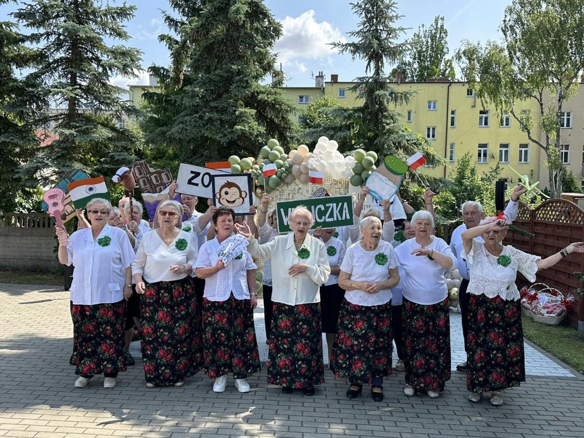 Grupa uczestników DDS, stoi na placu w rekach trzymaja różne tbliczki z napisami, rysunkami, flagami, w tle duże zielone choinki, blok, w tle 
