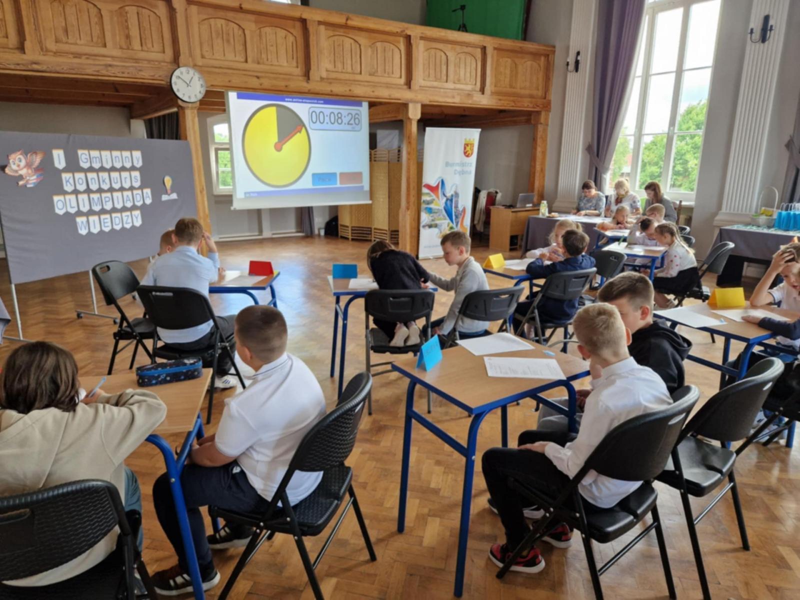 Uczniowie siedzą parami przy stolikach. w tle plansze. Aula.  