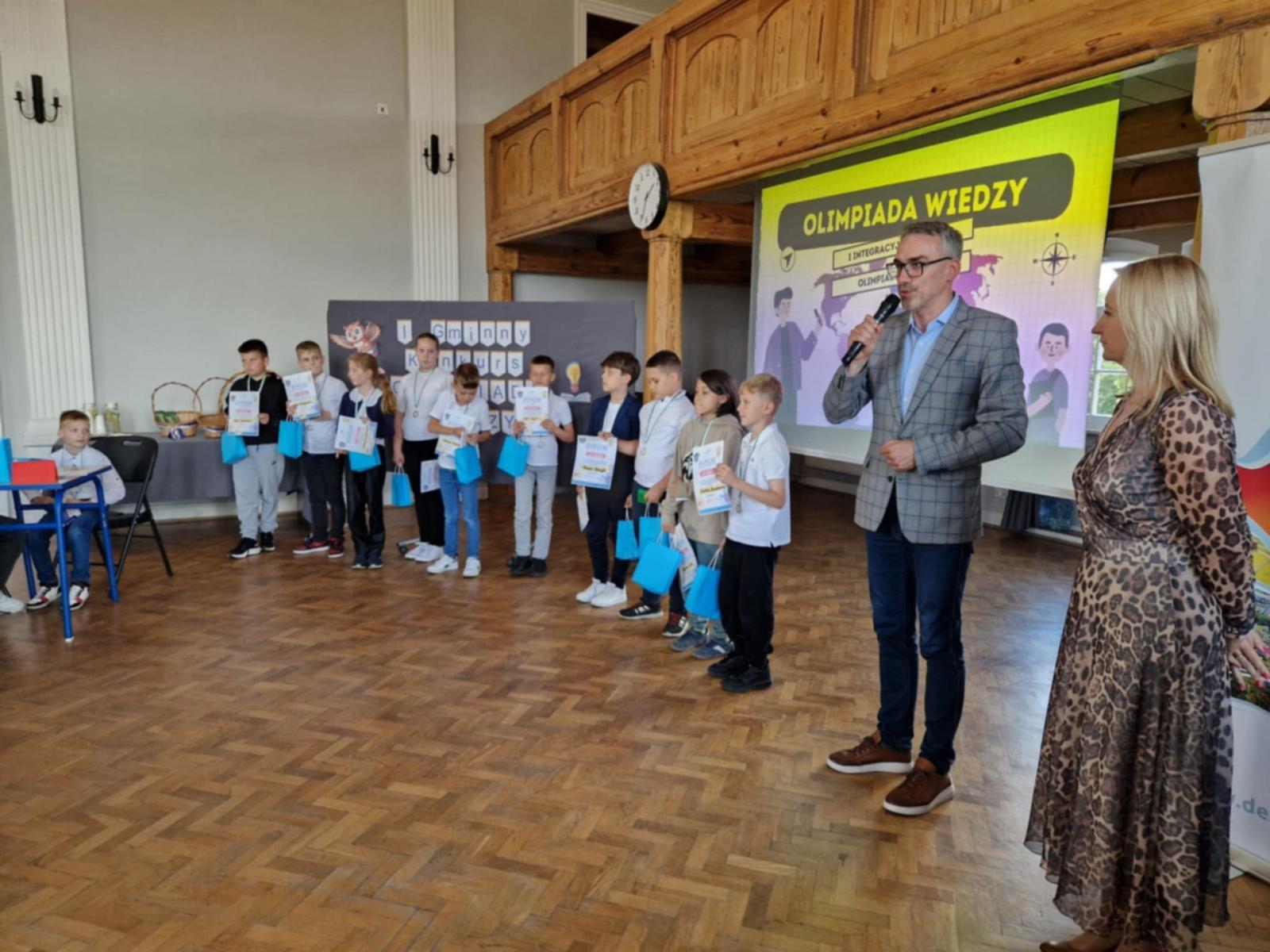 burmistrz Dębna stoi na auli. trzyma mikrofon w ręce. obok stoją uczniowie z dypomami i torebkami