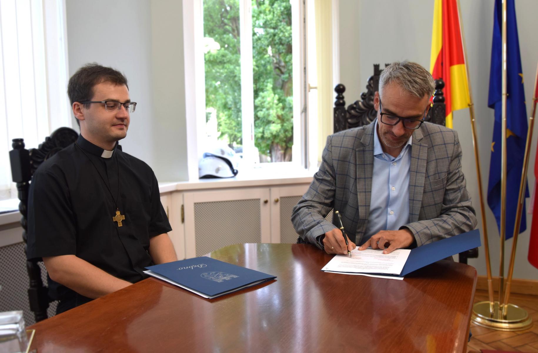 Przy stole siedzi dwóch mężczyzn, od prawej ksiądz, obok burmistrz podpisuje dokument