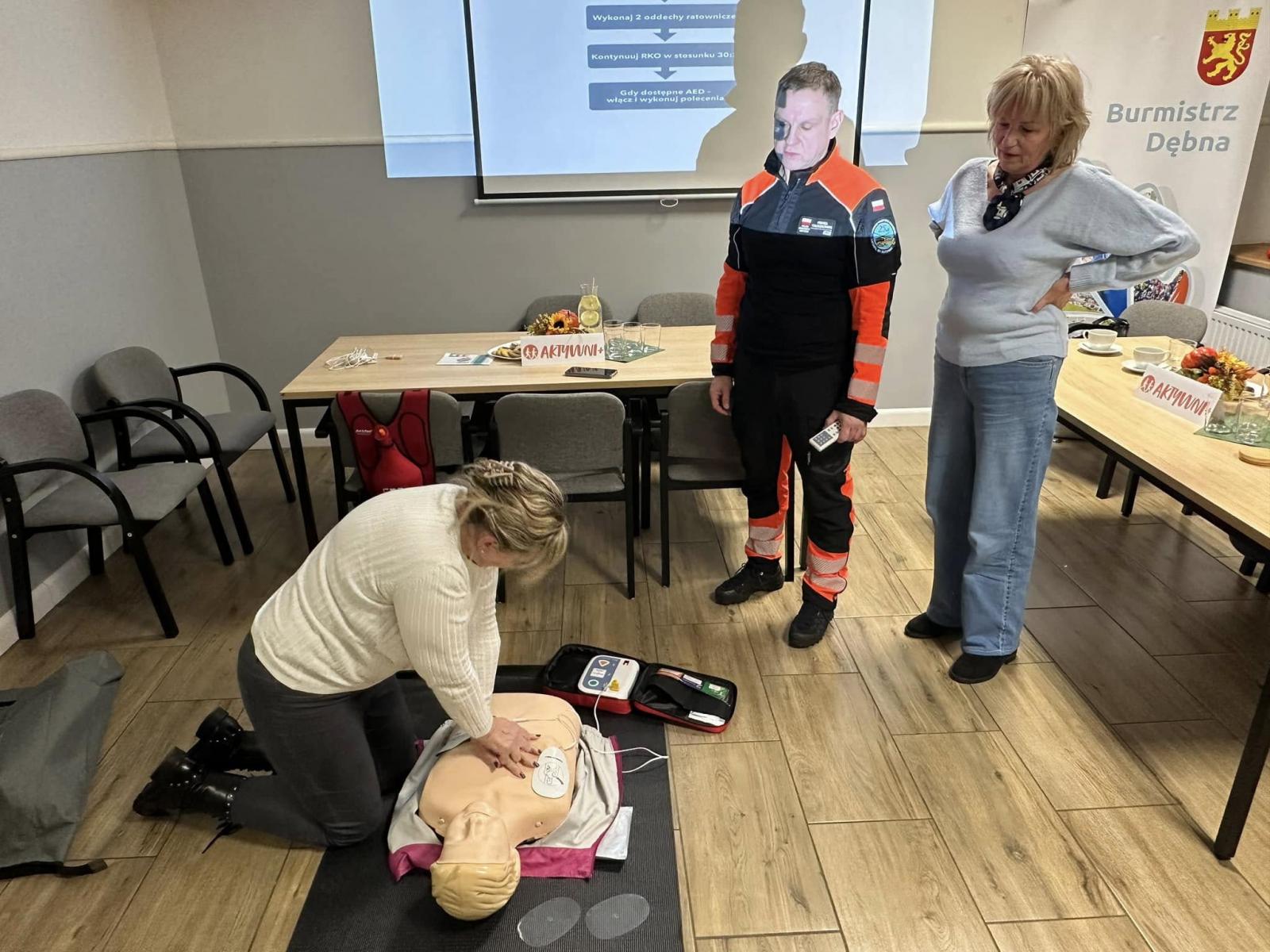 W sali, jedna kobieta kuca nad fantomem, jedna stoi i mężczyzna w stroju ratownika