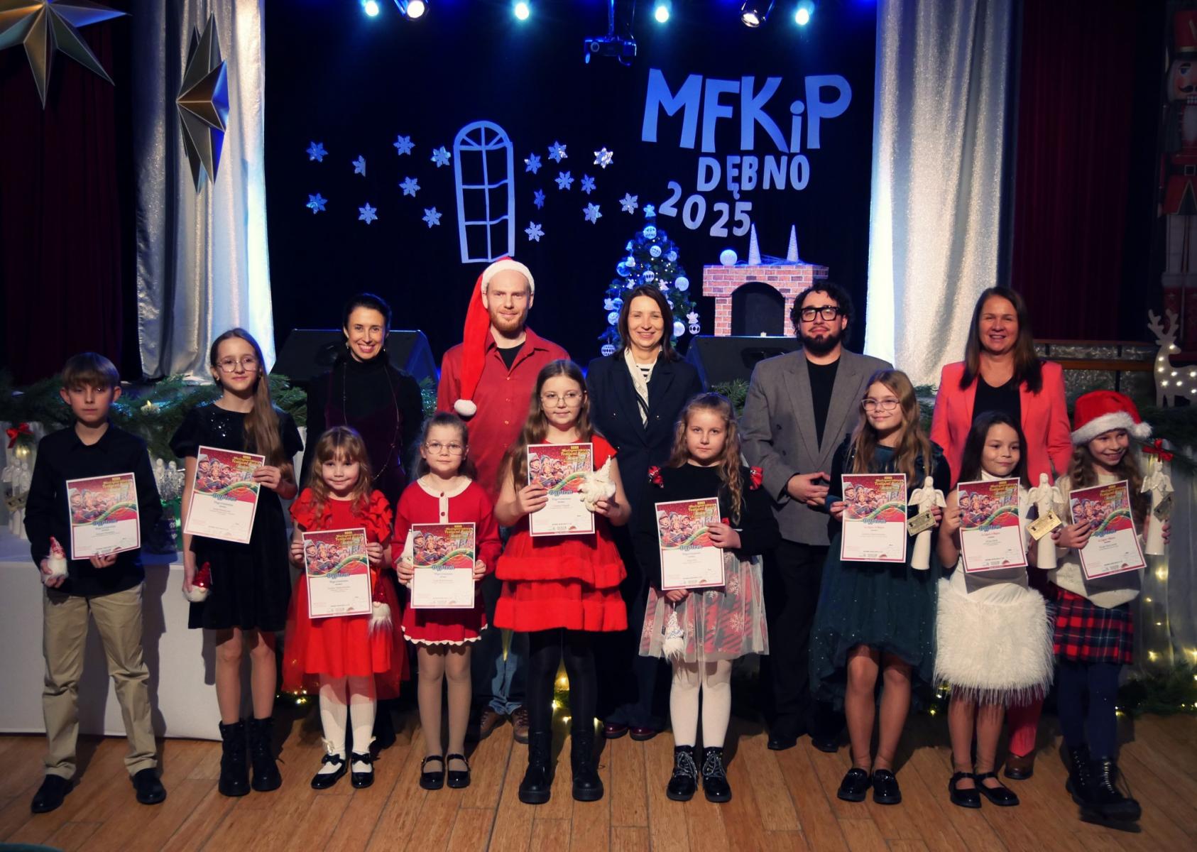 Grupa dzieci i dorosłych stoja w sali, nagrodzeni w pierwszym rzędzie trzymają dyplomy i białe anioły