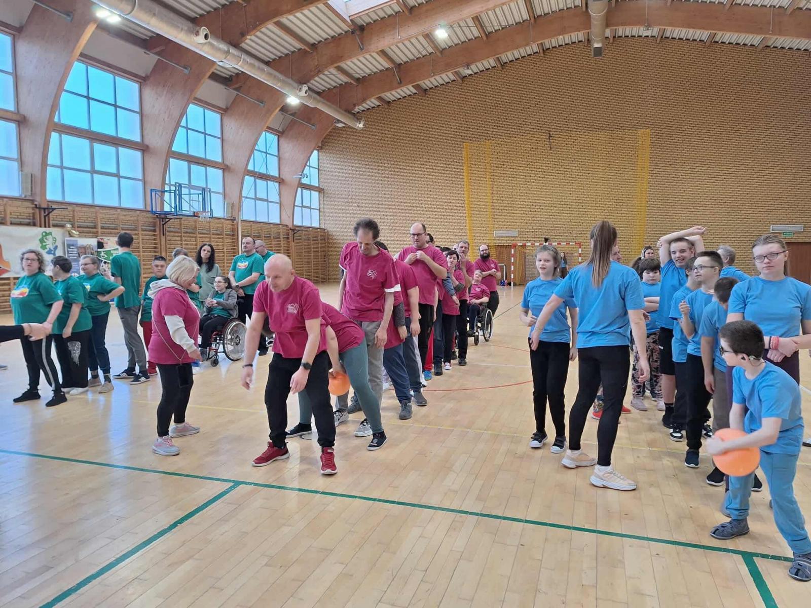 Uczestnicy podzieleni na trzy drużyny, każda w innych kolorach koszulek (zielone,r&oacute;żowe, niebieskie) stoją na hali w rzędach 