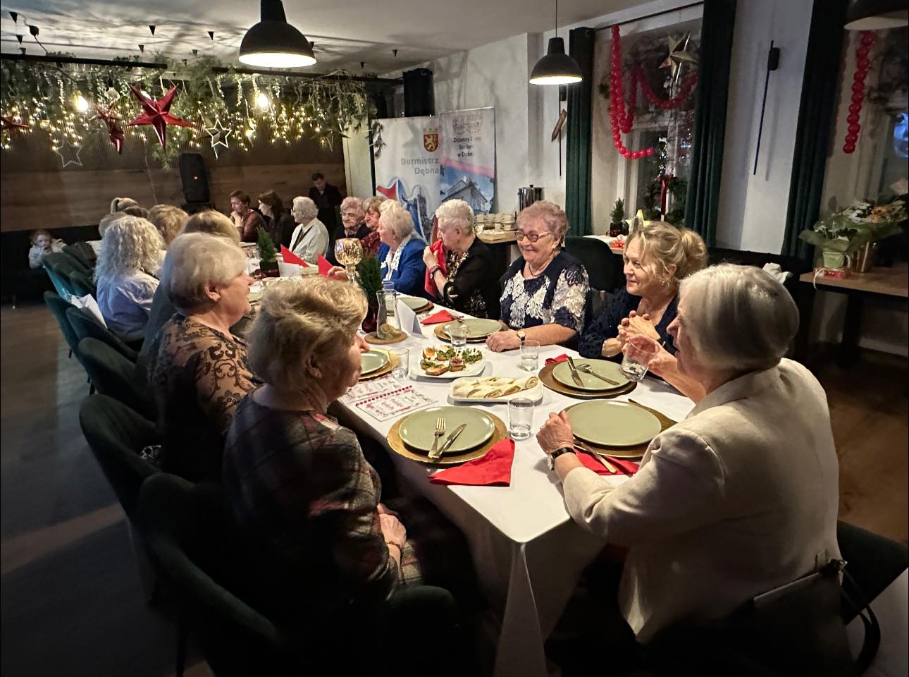 Kobiety siedzą przy wsp&oacute;lnym stole, odświętnie ubrane i sala przystrojona świątecznie 