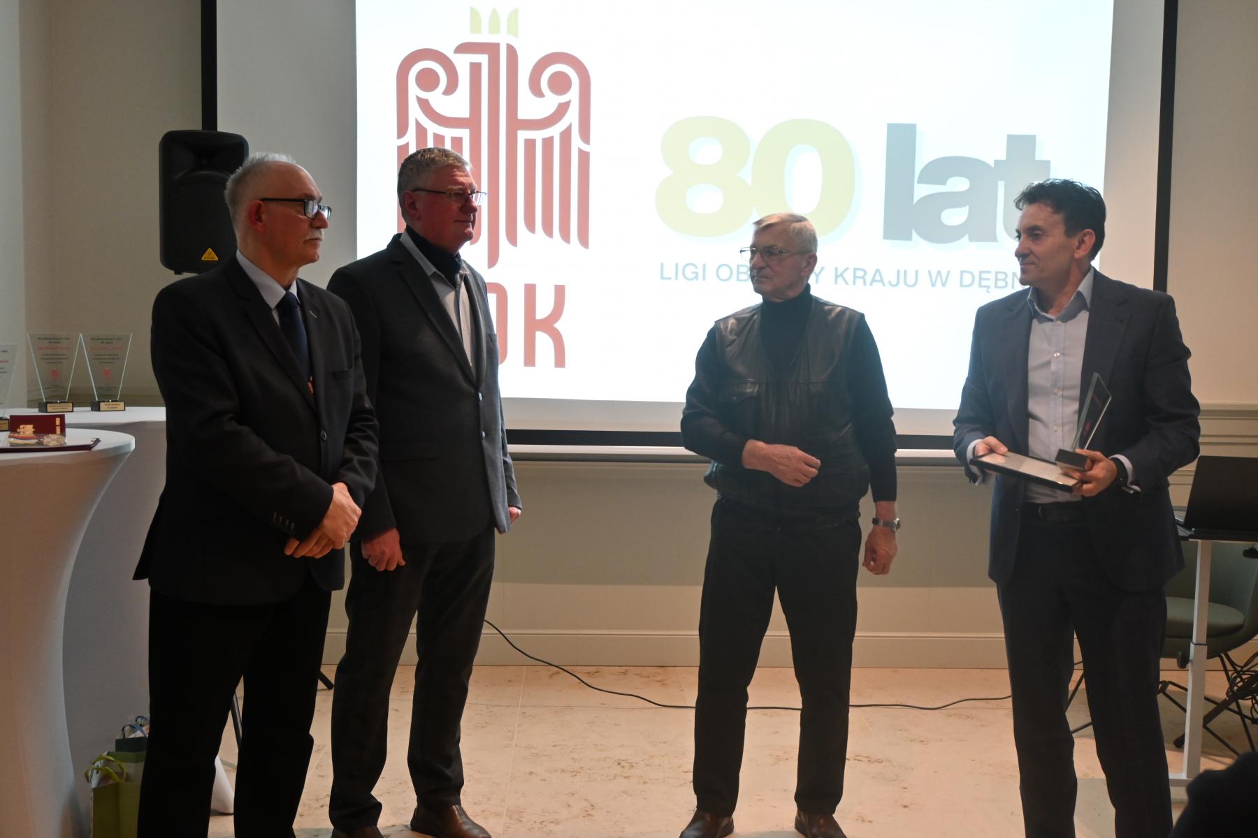 Czterech mężczyzn stoi w sali, w tle wyświetla się logo LOk i 80 lat