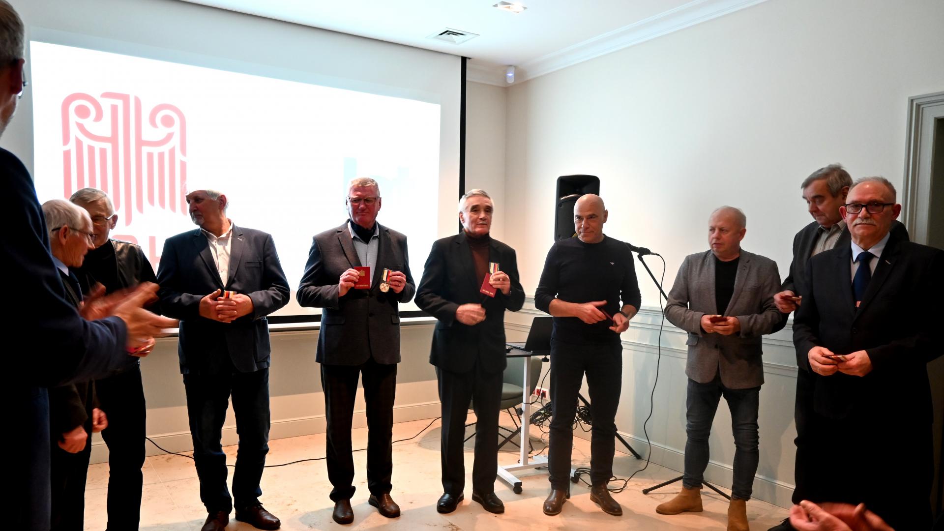 Grupa mężczyzn stoi w sali, w tle wyświetla się logo LOk 