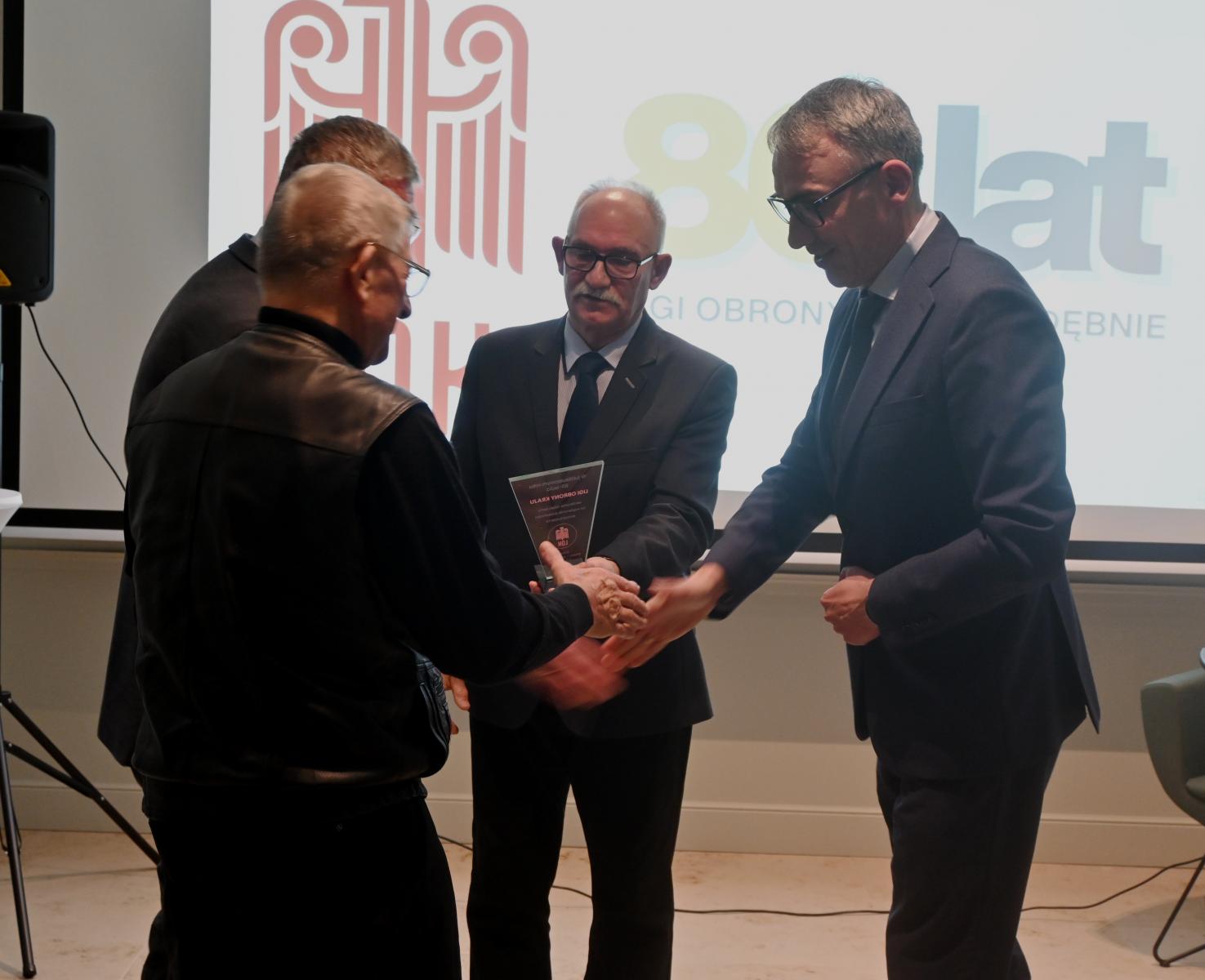 Czterech mężczyzn stoi w sali, w tle wyświetla się logo LOK i 80 lat