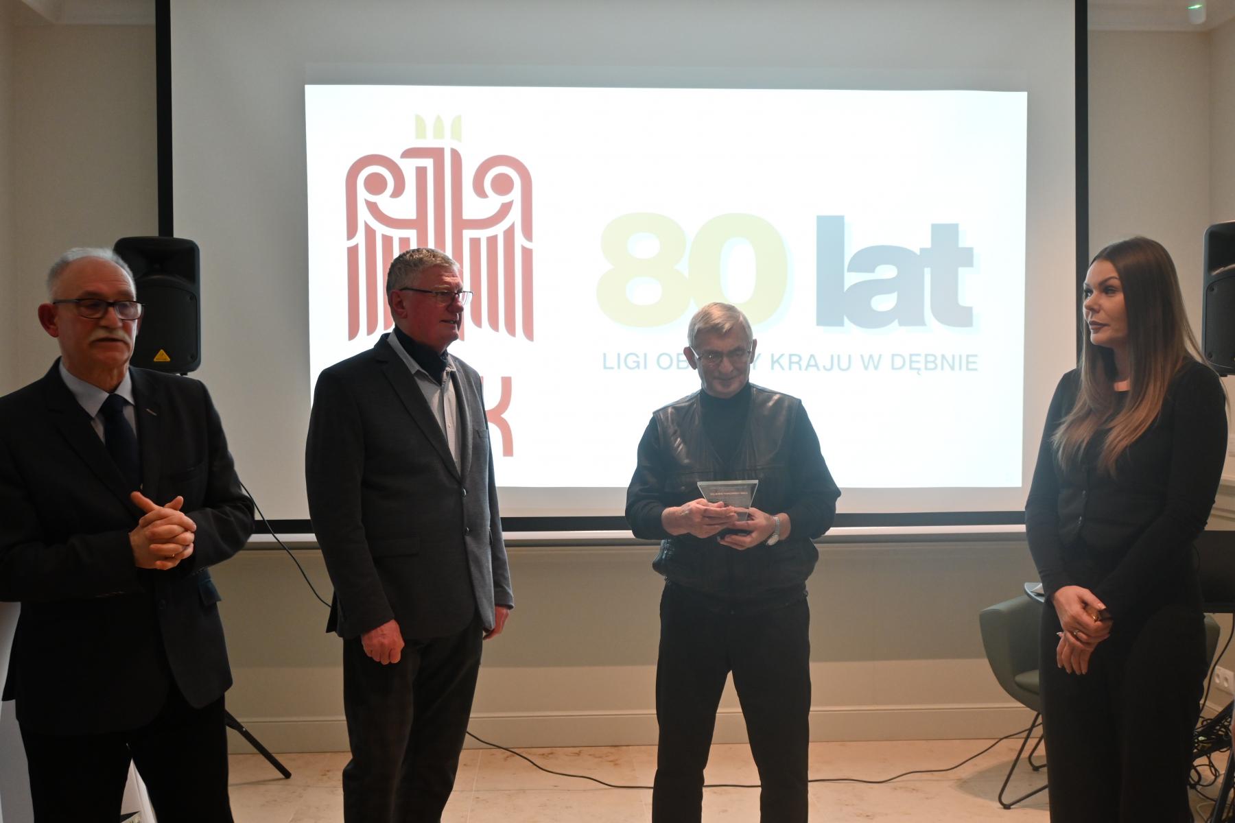 Trzech mężczyzn stoi w sali, od prawej stoi kobieta, w tle wyświetla się logo LOk i 80 lat
