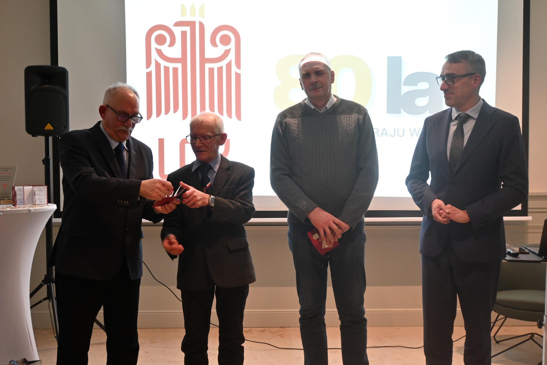 Czterech mężczyzn stoi w sali, w tle wyświetla się logo LOk i 80 lat