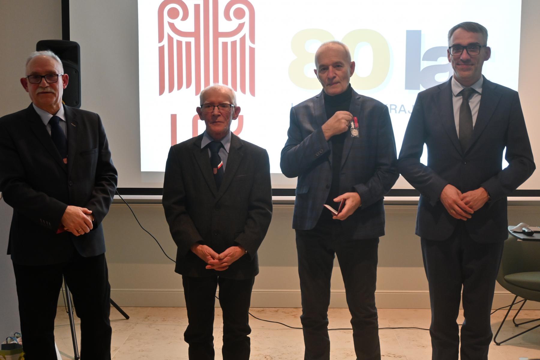 Czterech mężczyzn stoi w sali, w tle wyświetla się logo LOk i 80 lat