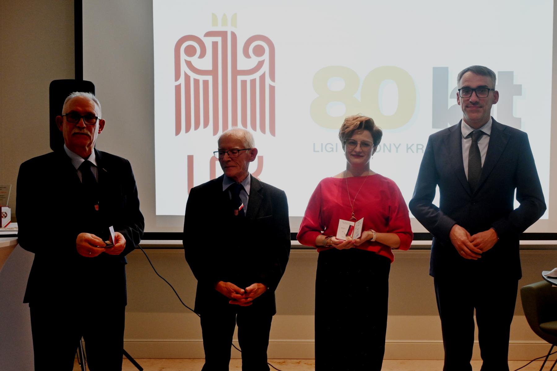 Od lewej stoją w sali: dw&oacute;ch mężczyzn, kobieta, mężczyzna, w tle wyświetla się logo LOk i 80 lat