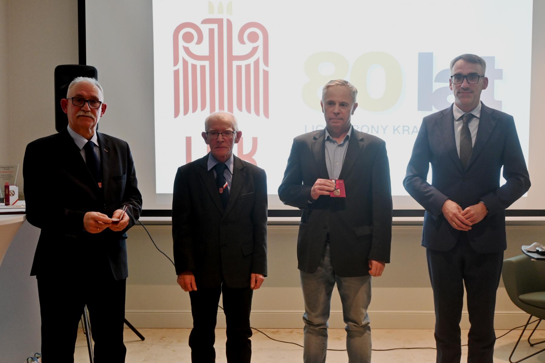 Czterech mężczyzn stoi w sali, w tle wyświetla się logo LOk i 80 lat