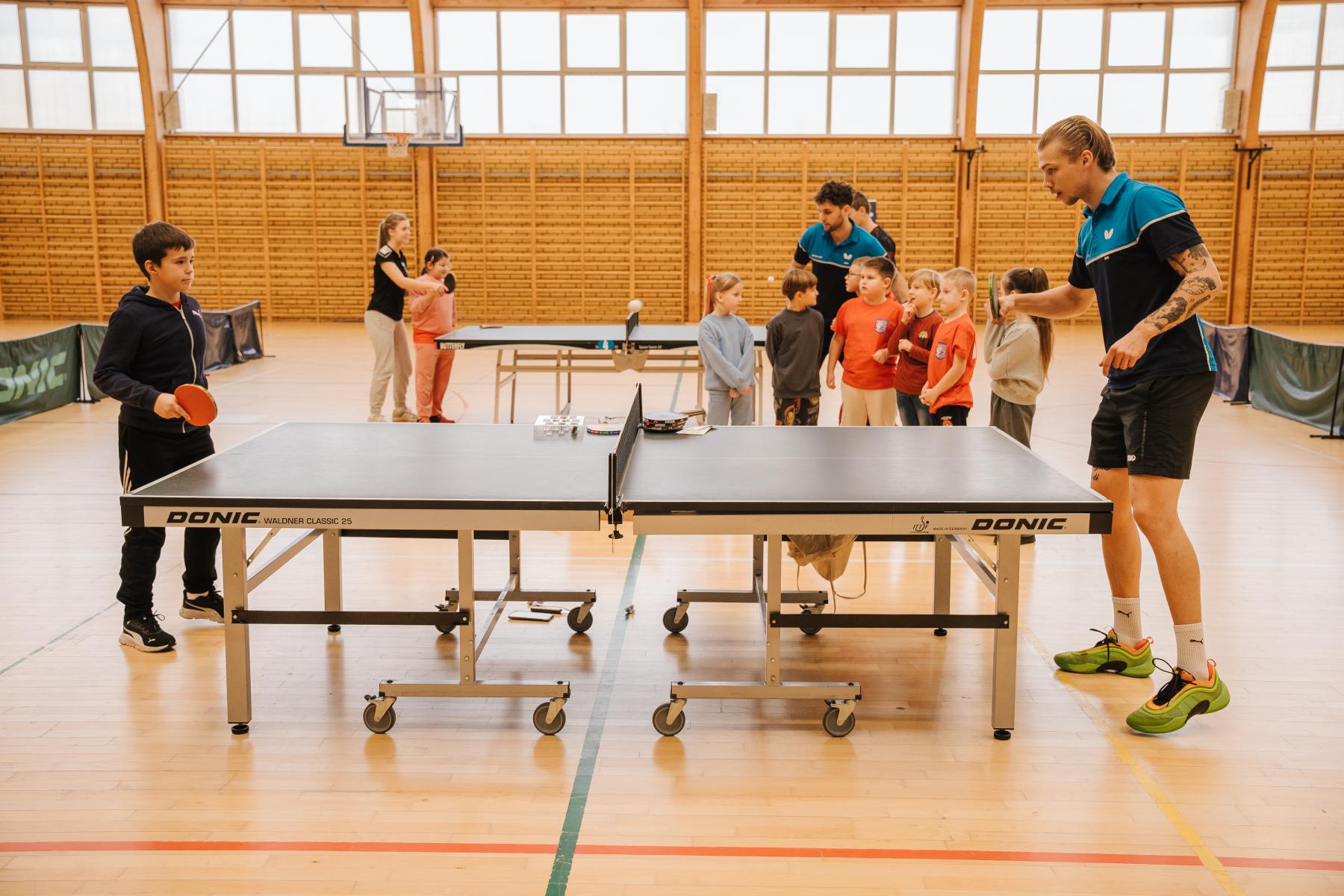 chłopczyk trzyma paletkę w ręku stoi przy stole do ping ponga, po drugiej stronie stoi młody mężczyzna, z boku grupka dzieci przygląda się 
