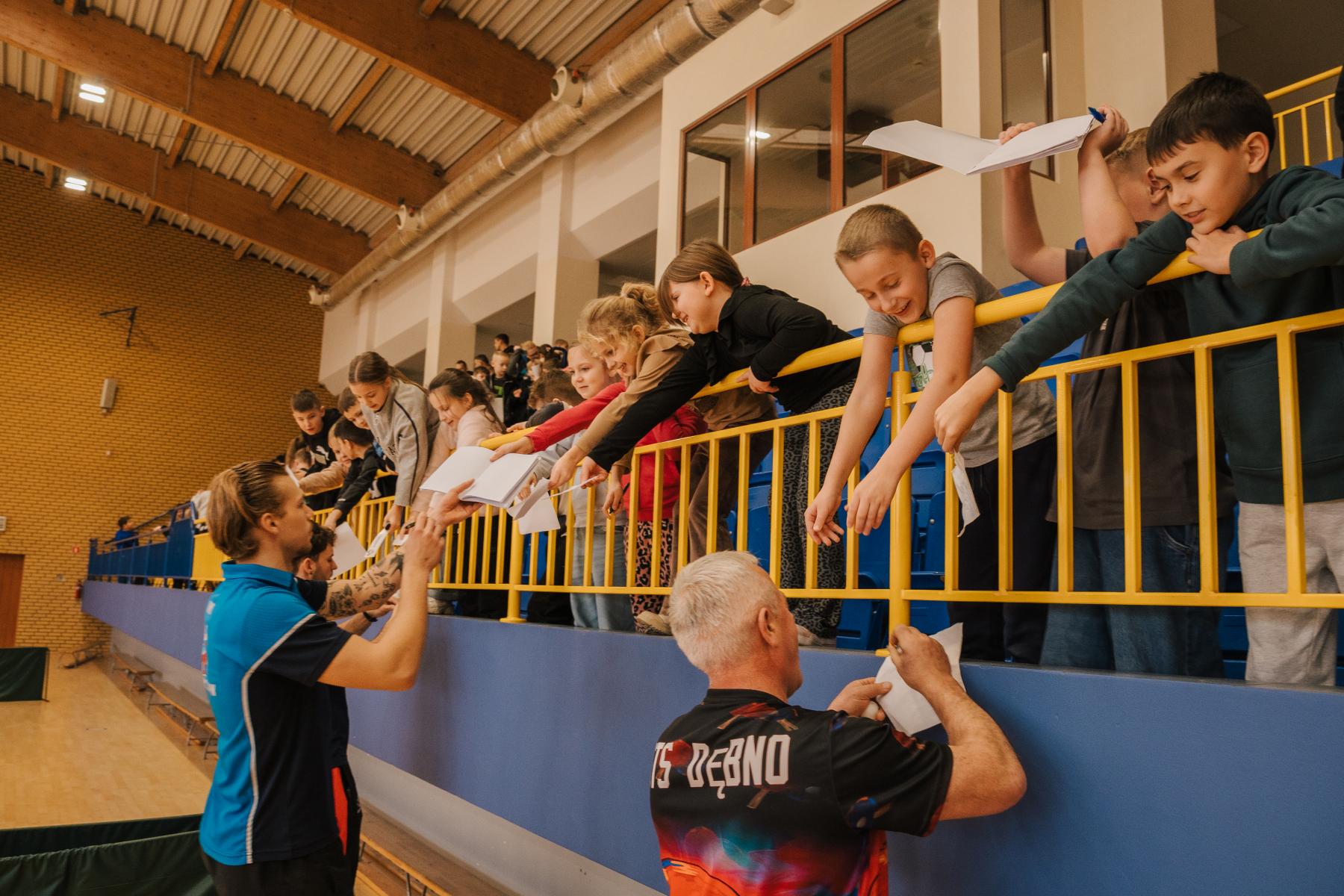 Ucnziwoie stoja na trybunach, niżej dwóch mężczyzn podpisują sie na kartkach 