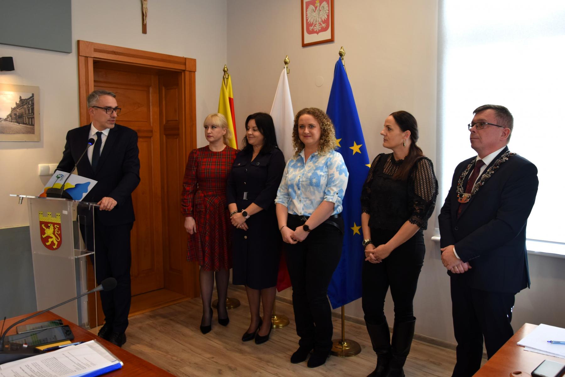 W sali posiedzeń burmistrz stoi przy m&oacute;wnicy, obok stoją cztery kobiety i mężczyzna. W tle na ścianie godło i flagi na stojakach