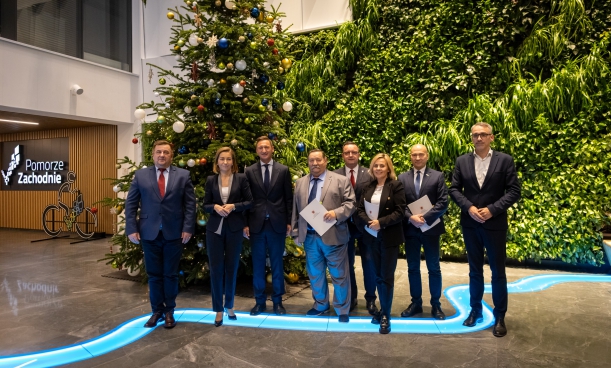 W wysokiej auli, na tle dużej choinki z bombkami stoi 8 osób.