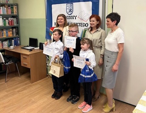 Trójka dzieci stoi trzyma dyplomy w rękach, za nimi trójka dorosłych kobiet