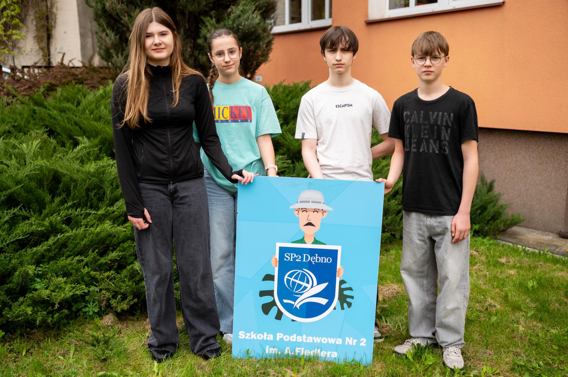 Od lewej dwie uczennice, dwóch chłopców, przed nimi tarcza szkoły, którą trzymają. Stoją na zewnątrz, w tle zielone krzewy i budynek szkoły