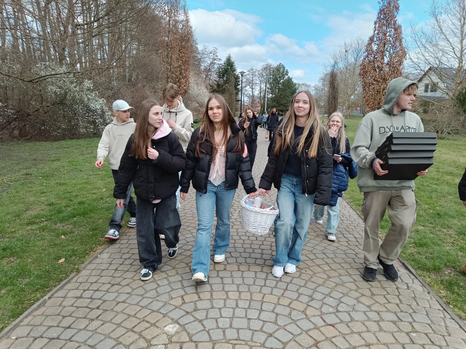 Promenadą idzie grupa uczniów, w pierwszej lini trzy dziewczyny, dwie z nich niosą biały koszyk  i chłopak