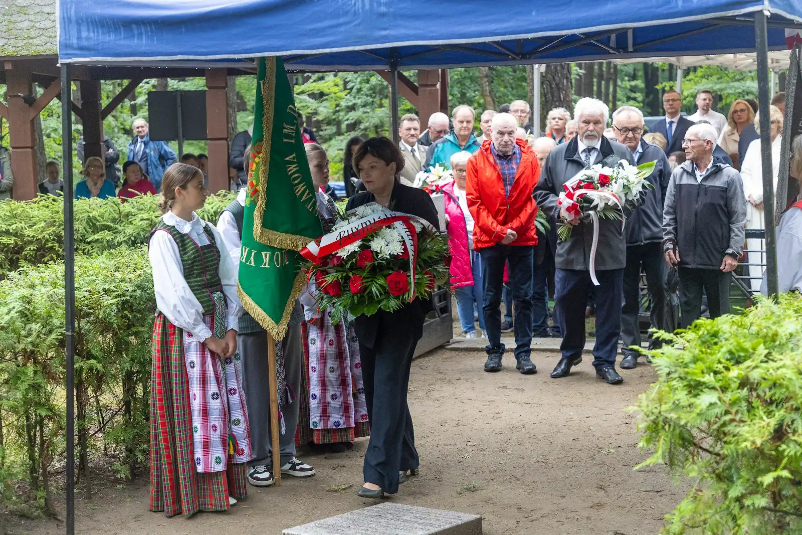 Pani sekretarz idzie z wieńcem, pod niebieskim namiotem. Za nią stoją kolejni ludzie z kwiatami