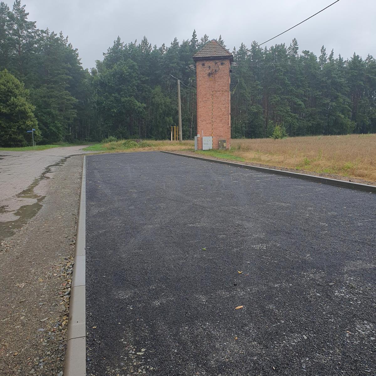 Wydzielony obszar z z masy asfaltowej, w oddali budynek wieżyczka, las  