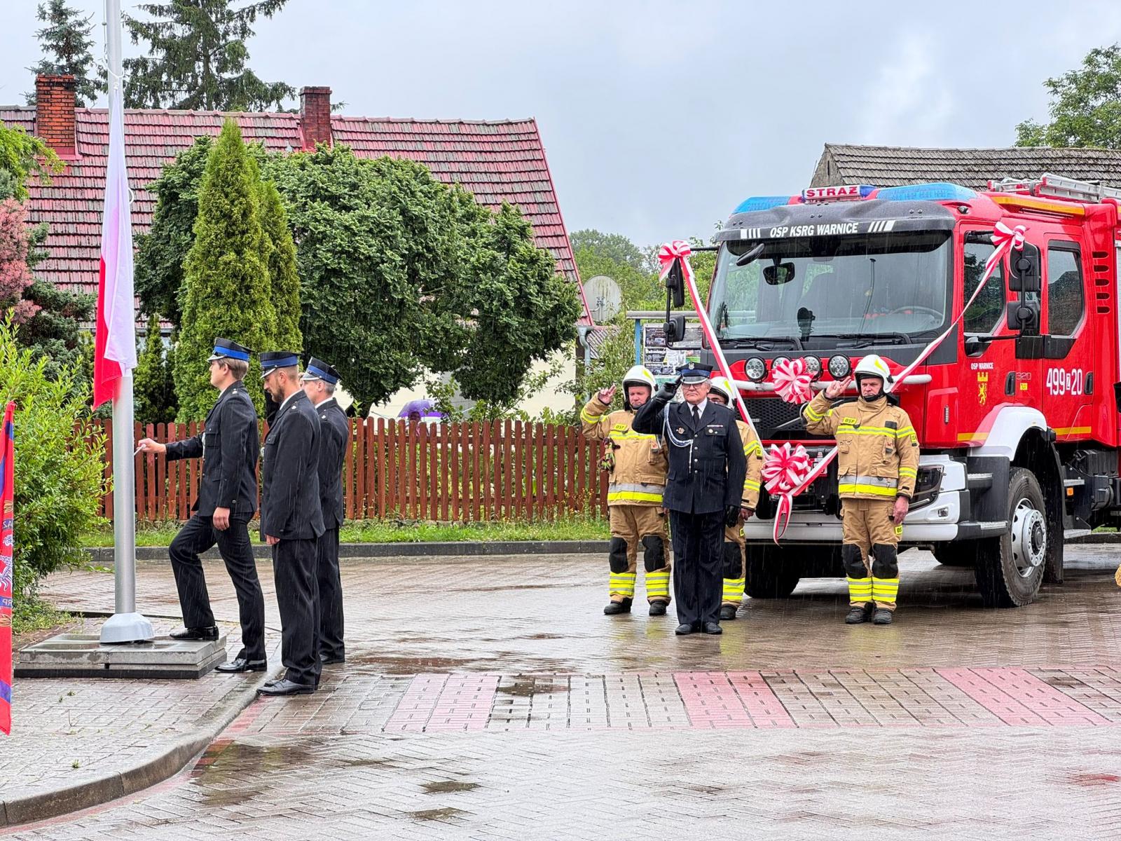 Strażacy wciągają biało-czerwoną flagę na maszt. w tle wóz strażacki z kokardami. przed nim salutuje trzech strażaków w mundurach