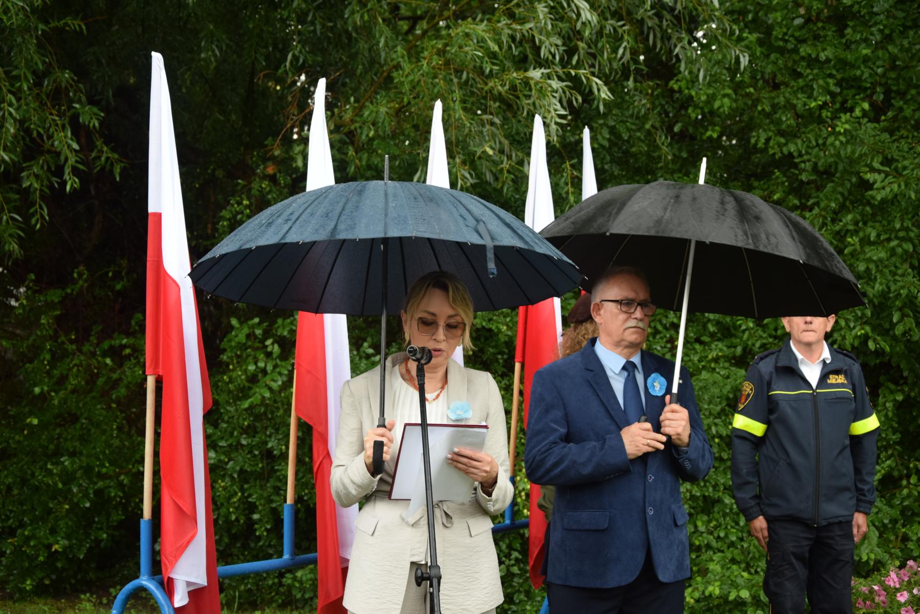 Przewodnicząca Stowaryszenie Kresy stoi pod parasolem, przed mikrofonem, za nią stoi mężczyzna z parasolem w tle flagi Polski