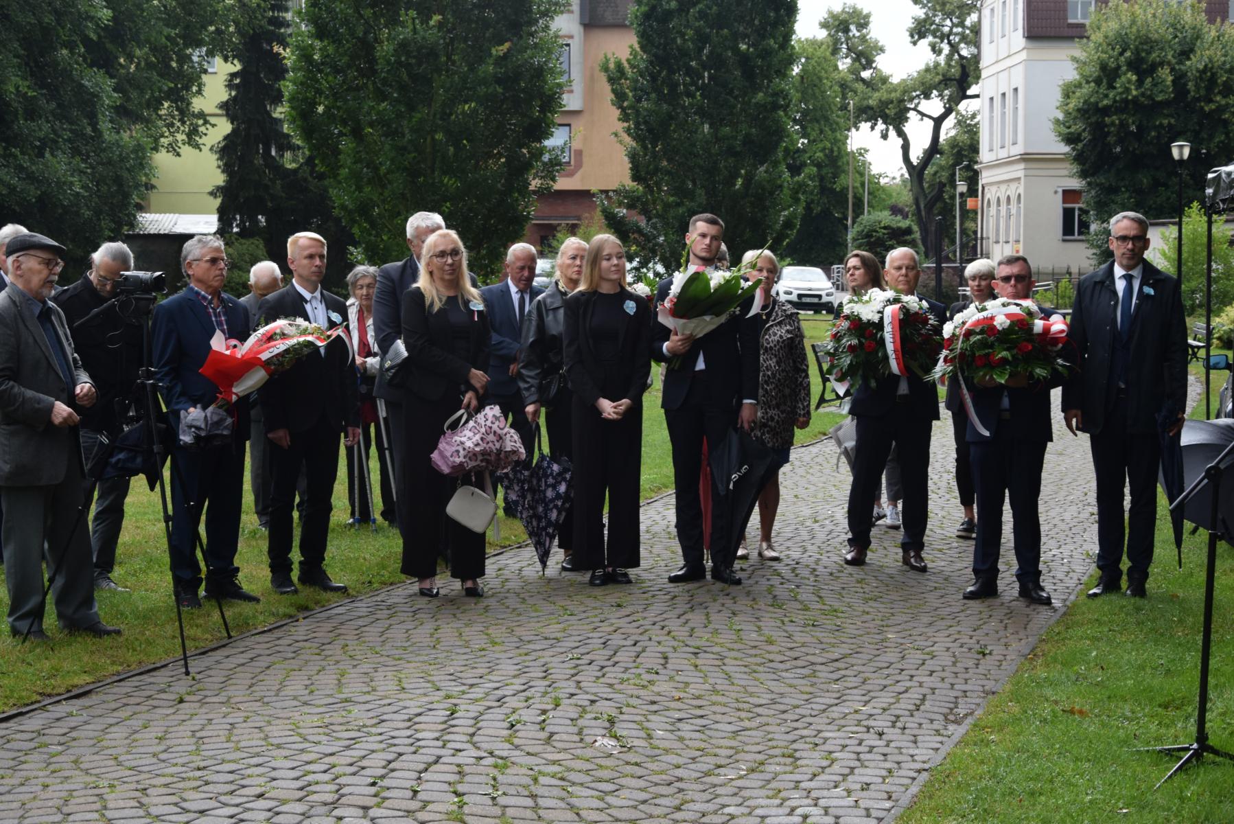 Uczestnicy uroczystości stoją na promenadzie. Trzymają wiązanki biało-czerownych kwiatów. 