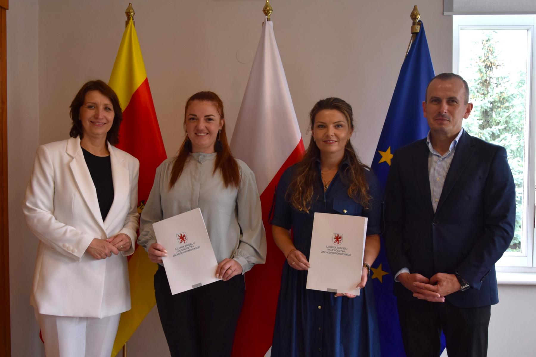 W sali od lewej stoją: trzy kobiety i mężczyzna. W tle ściana i trzy flagi: gminna żółto-czerwona, Polski biało-czerwona, UE