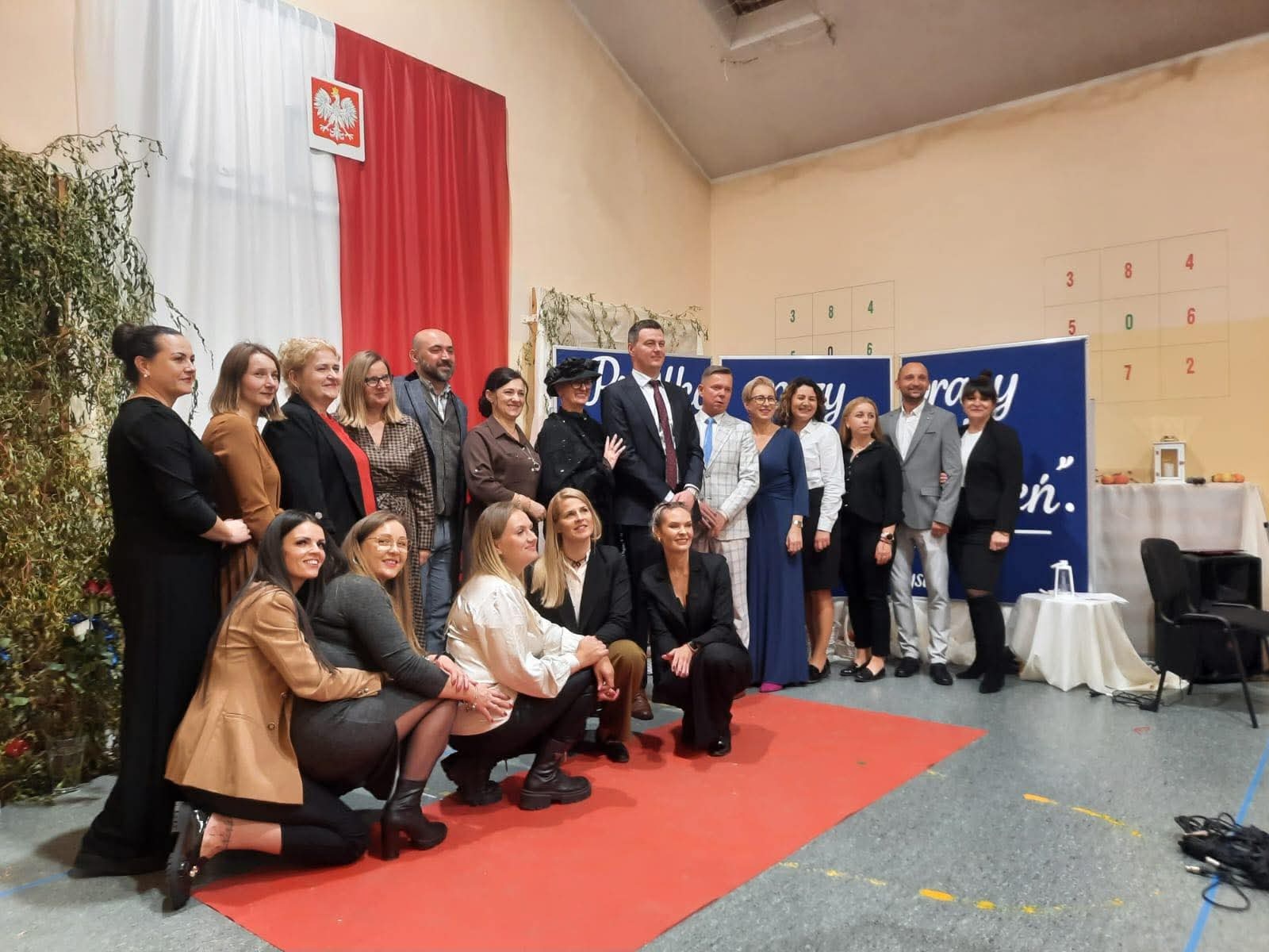 Grupa kadry nauczycielskiej stoi w sali, w tle biało -czerwone płótno w pionie na nim godło Polski 