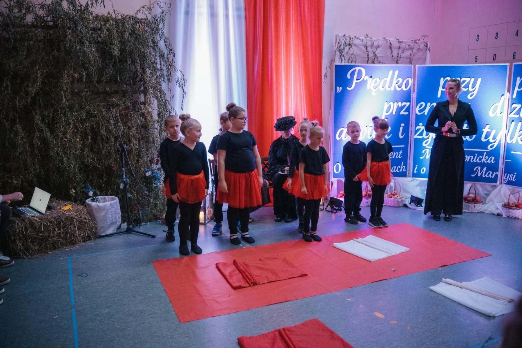 Grupa dziewczynek ubrane w czerowne spódniczki i czarne koszulki, obok po prawej stronie stoi kobieta. Przed nimi czerwone płótno rozłożone na ziemi. 