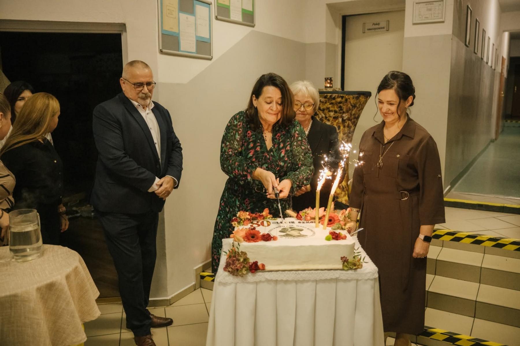 Od lewej stoją: mężczyzna, kobieta kroi tort, kobieta stoi. 