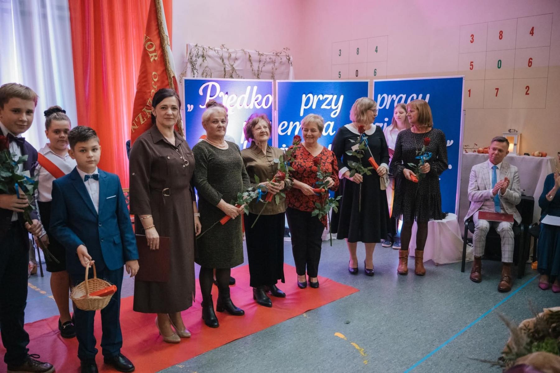 Od lewej w sali stoją: dwóch ucznió, dyrektor szkoły, pięć kobiet trzymają kwiat w dłoni