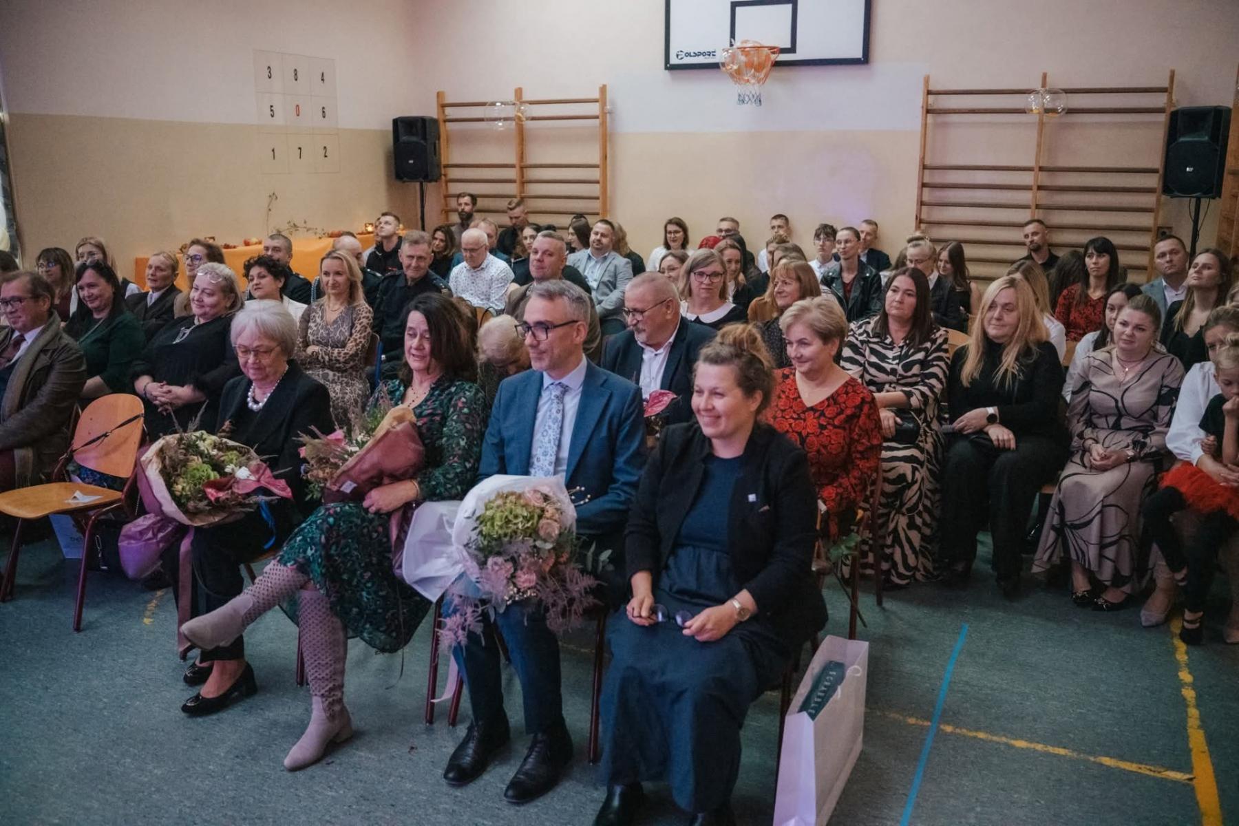 Goście siedzą na krzesełkach  na sali gimnastycznej 