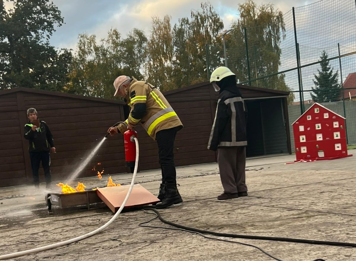 Mężczyzna w kurtce strażaka gasi pożar, za nim stoi dziecko ubrane w str&oacute;j strażacki i kask 
