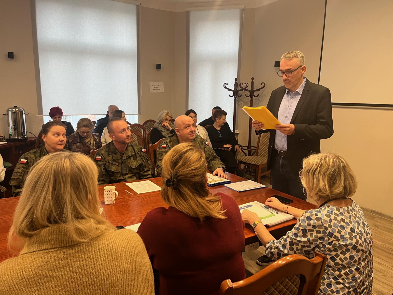 Burmistrz Debna stoi z dokumentem w ręku, przy stole siedzi trzech wojksowych w mundurach i pracownicy urzedu