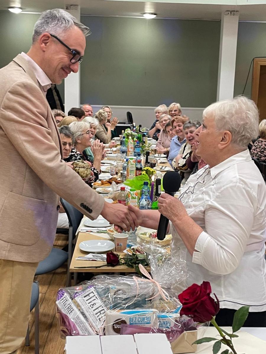 Burmistrz Dębna uśmiechnięty ściska dłoń pani Jaworskiej. Za nimi w tle siedzą ludzie przy stoliku
