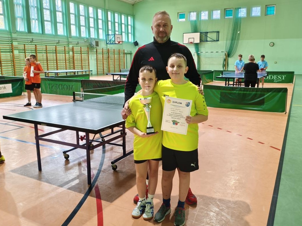 Dwóch chłopców w zielonych koszulkach, za nimi mężczyzna stoją w sali sportowej