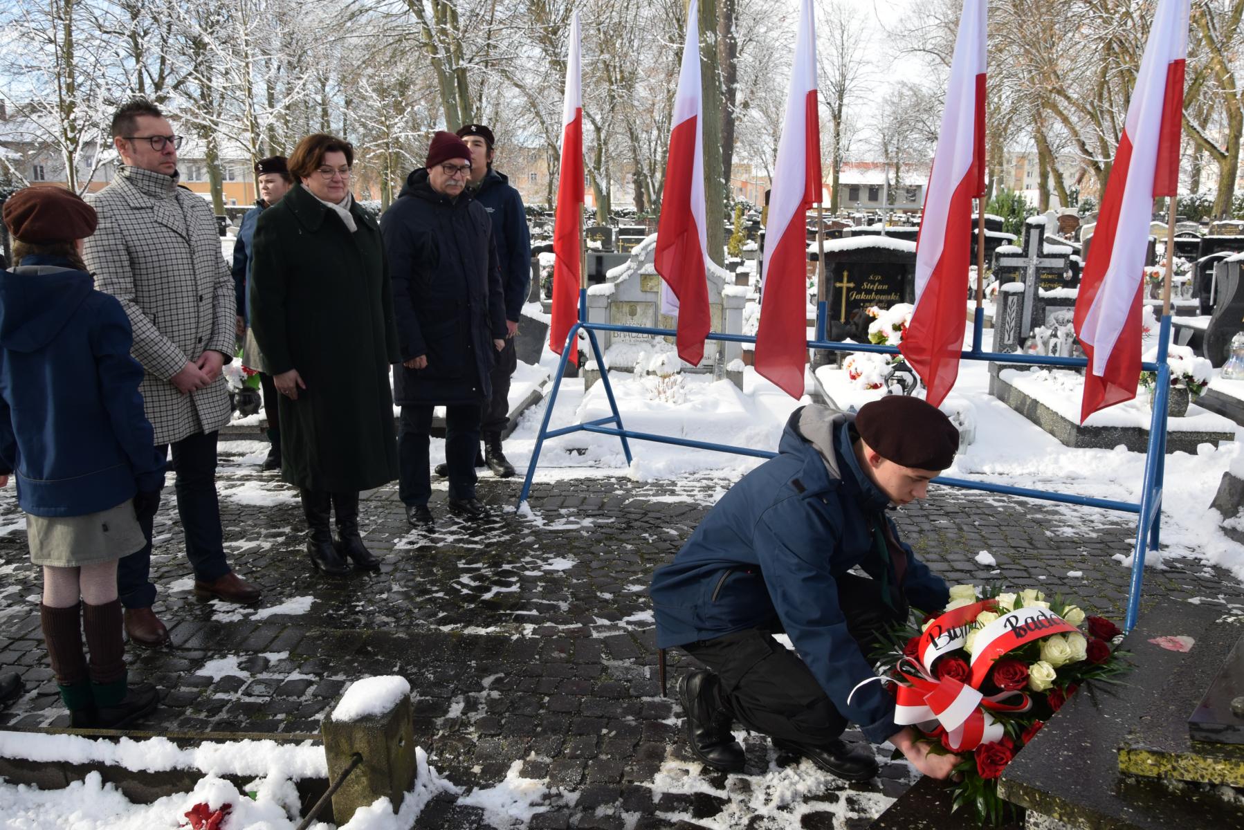 Druh składa kwiaty na grobie, za nim stoi delegacja władz samorządowych