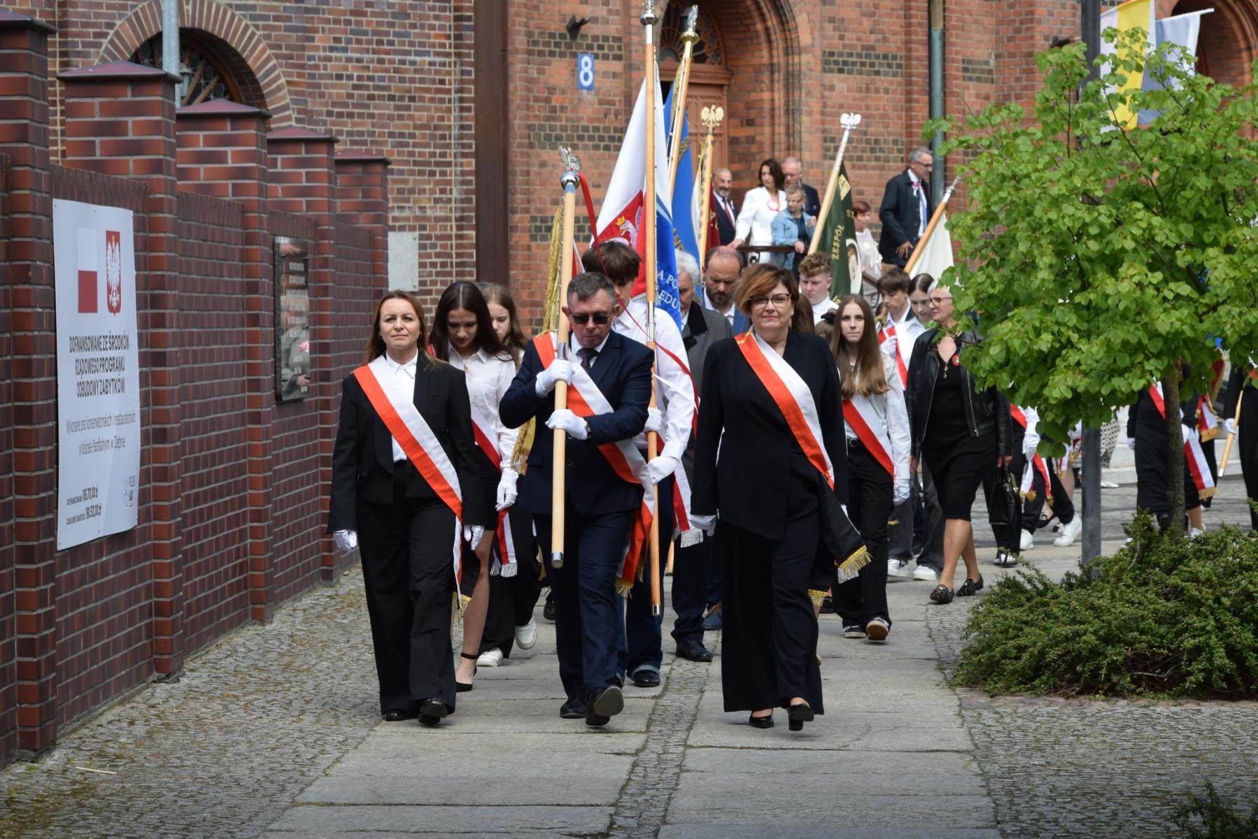 Poczty sztandarowe maszerują. w tle ludzie wychodzą z koscioła. po lewej mur 
